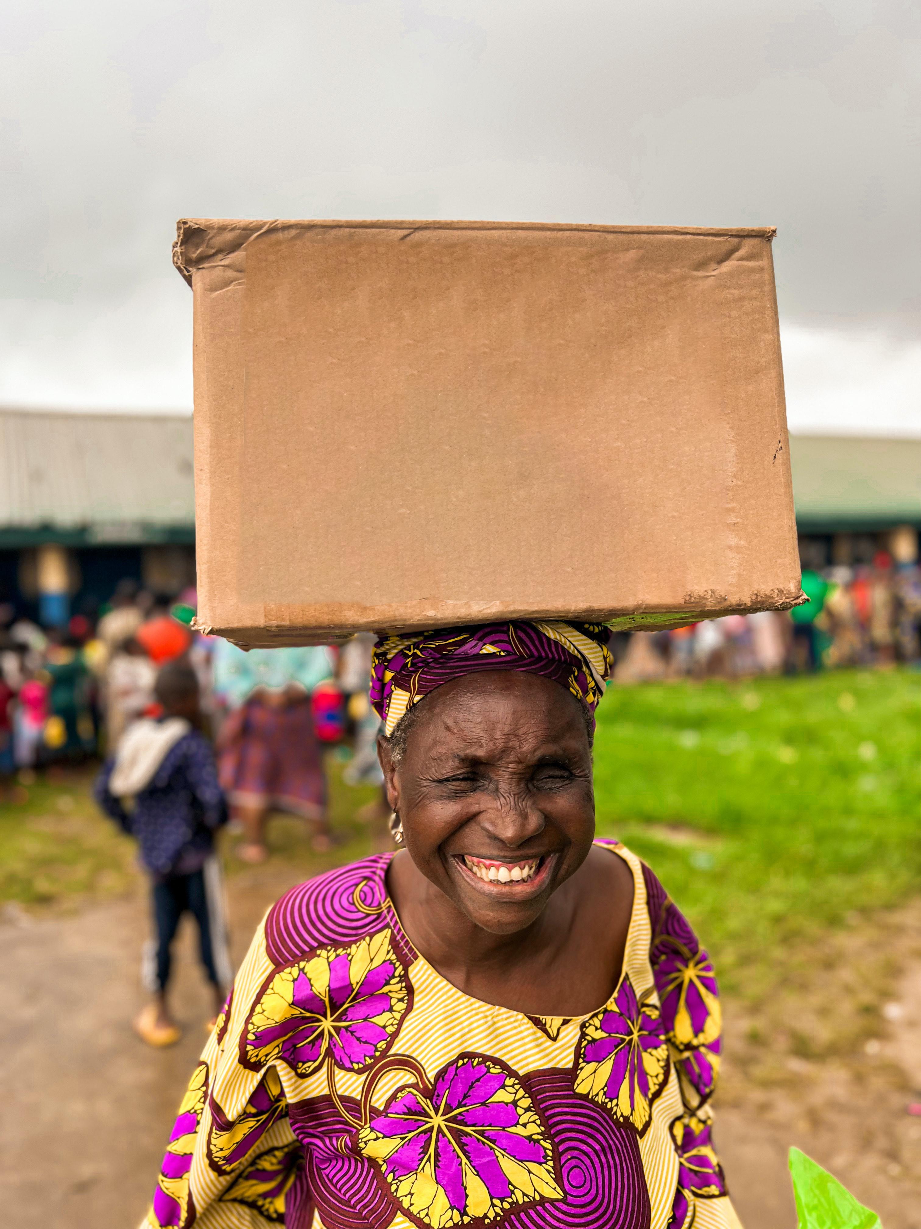 an old lady smiling and carrying a brown carton on her head